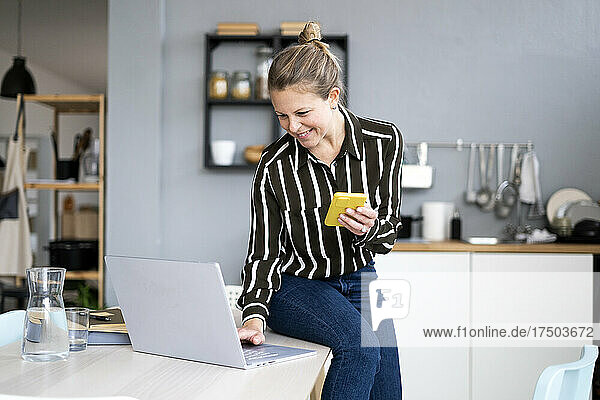 Smiling freelancer working on laptop holding mobile phone in kitchen