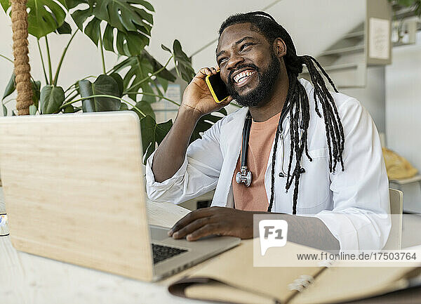 Happy hipster doctor talking on mobile phone sitting with laptop at home office