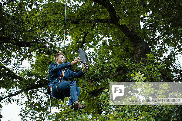 Smiling businessman tied up with rope on tree doing video call through tablet PC