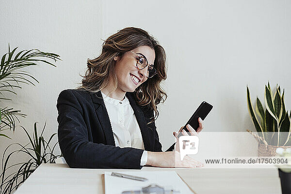 Smiling businesswoman with eyeglasses using smart phone at office