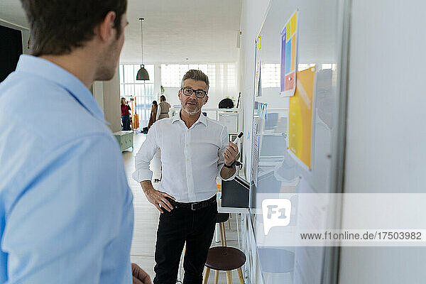 Businessman with hand on hip explaining strategy to colleague on white board at office