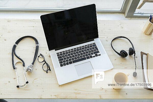 Laptop inmitten von Stethoskop und Tasse mit Kopfhörern auf dem Schreibtisch im Heimbüro