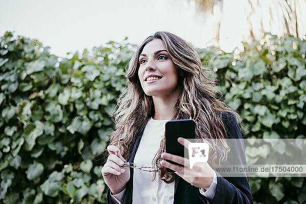 Young businesswoman holding eyeglasses and smart phone near hedge