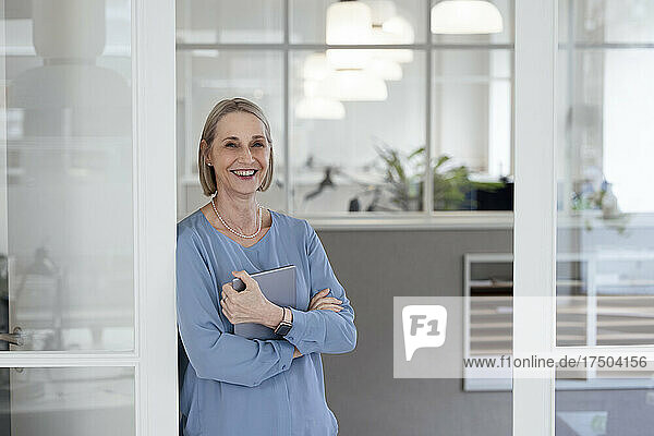 Smiling businesswoman with tablet PC leaning on office door