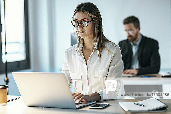 Junge Geschäftsfrau mit Brille arbeitet am Laptop mit Kollegin im Hintergrund im Büro