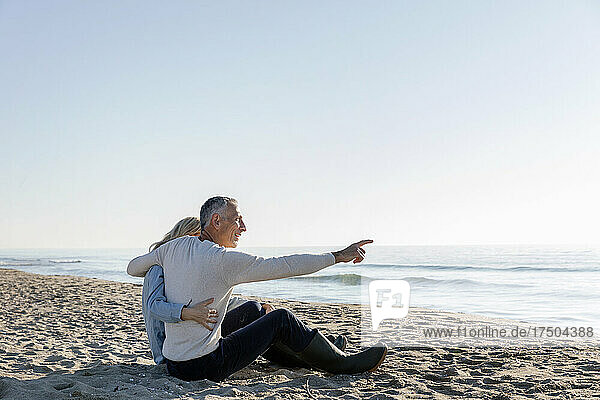 Mann zeigt aufs Meer und sitzt mit Arm um Frau am Strand