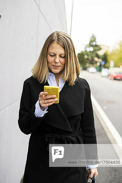 Smiling woman texting through smart phone walking by wall