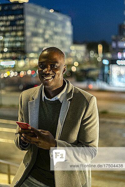 Businessman with smart phone leaning on railing in city at night