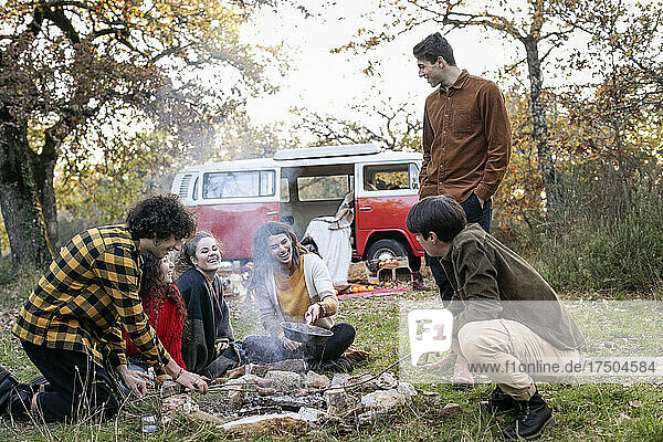 Happy friends cooking sausages in autumn forest on picnic