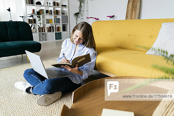 Smiling woman with laptop writing in book at home