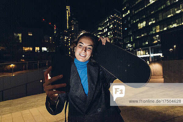 Smiling businesswoman with skateboard video calling through smart phone at night in city