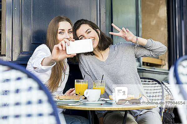 Junge Frau macht ein Selfie mit einer Freundin und zeigt im Café ein Friedenszeichen