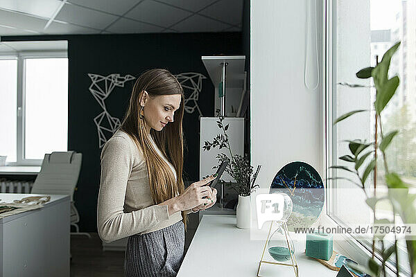 Saleswoman using smart phone near window sill in travel agency