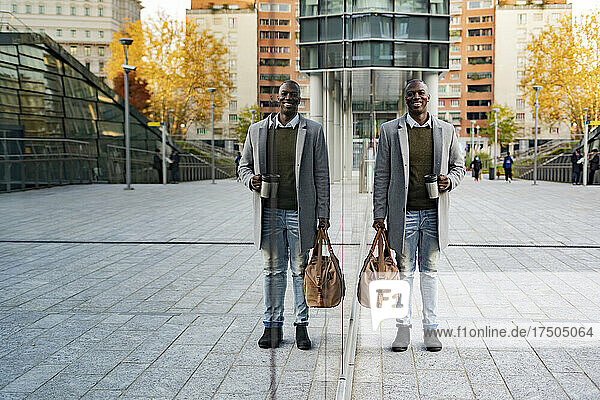 Businessman with duffel bag standing by reflection on glass wall