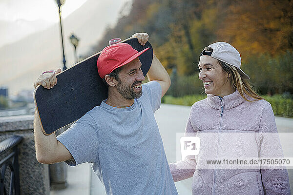 Smiling woman talking to boyfriend holding skateboard on footpath