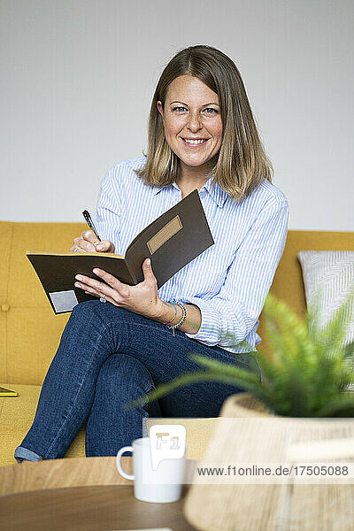 Smiling woman sitting on sofa writing in book at home