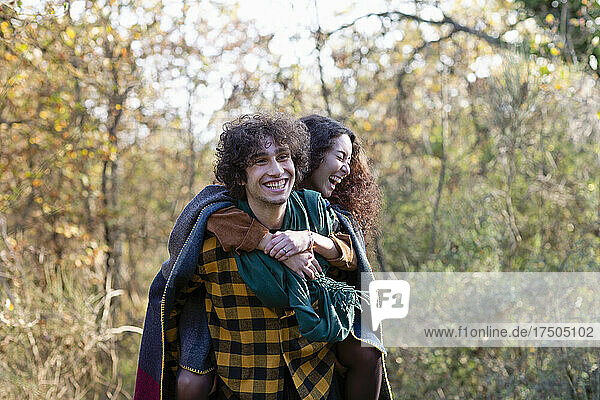 Cheerful man giving piggyback to girlfriend laughing in autumn forest