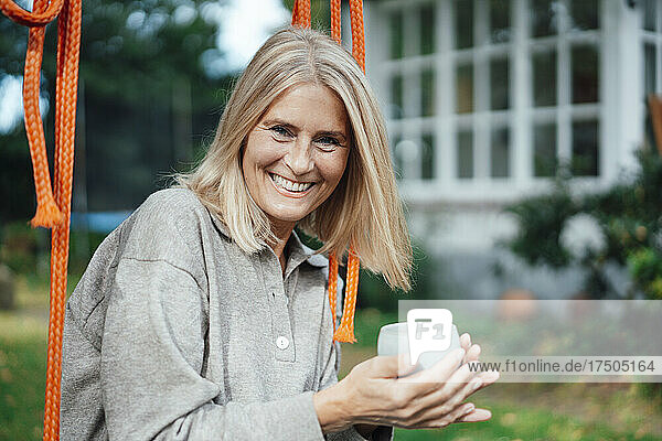 Happy woman holding coffee cup on swing at backyard