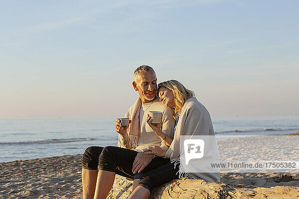 Ein Paar sitzt bei Sonnenuntergang auf einem Baumstamm am Strand und trinkt Tee