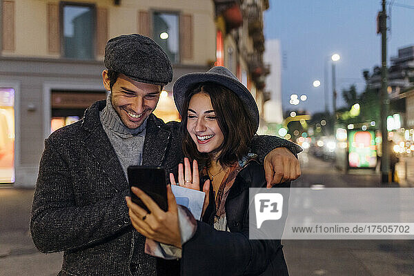 Happy young couple waving on video call through smart phone in city