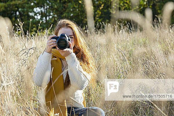 Woman photographing through camera while sitting in grass on sunny day