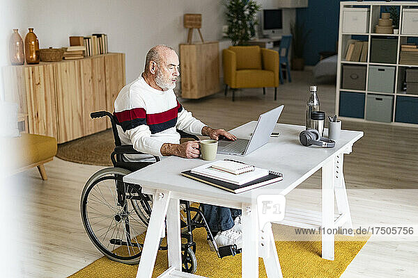 Leitender Geschäftsmann im Rollstuhl mit Kaffeetasse und Laptop im Heimbüro