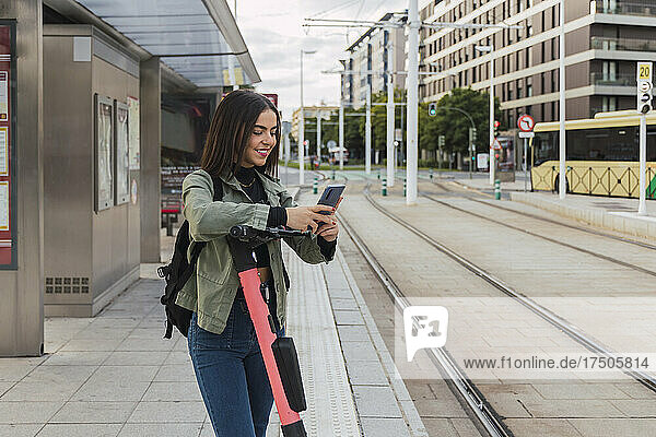 Frau mit Elektroroller benutzt Mobiltelefon an Straßenbahnhaltestelle
