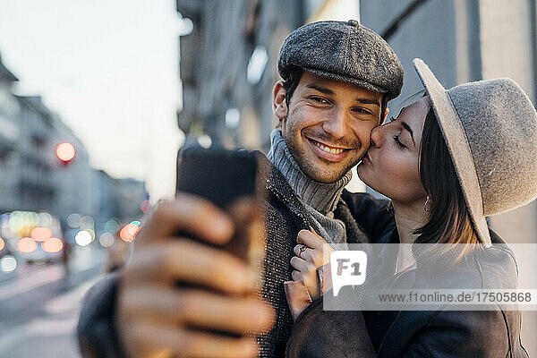 Junge Frau küsst ihren glücklichen Freund  während sie ein Selfie mit dem Smartphone macht