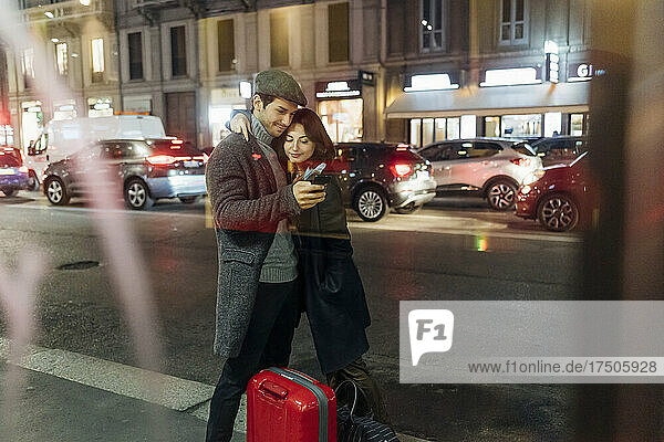 Young couple with wheeled luggage using smart phone on city street