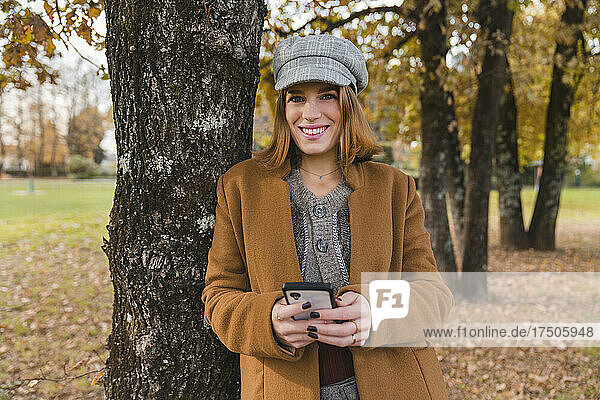 Smiling woman with smart phone leaning on tree trunk