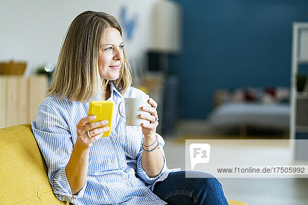 Thoughtful woman holding coffee cup and mobile phone at home