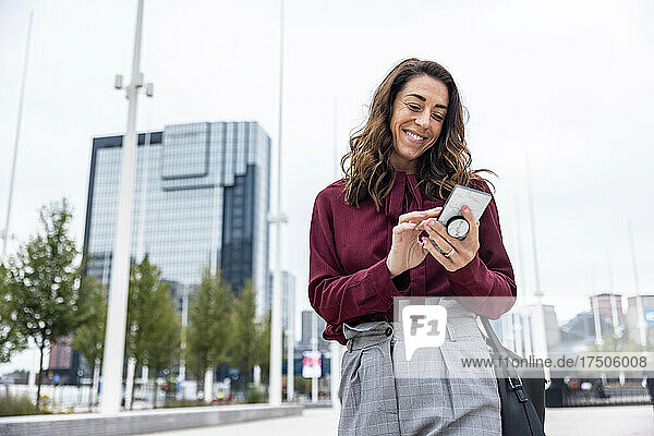 Lächelnde Geschäftsfrau  die mit ihrem Smartphone auf der Straße in der Stadt unterwegs ist