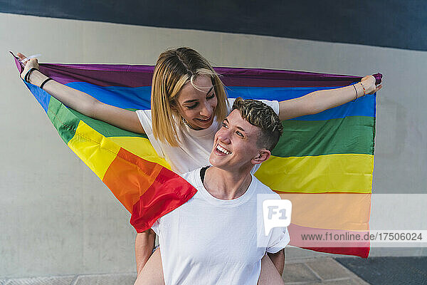 Happy woman piggybacking girlfriend with rainbow flag in front of wall