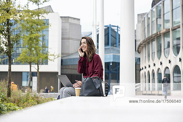 Lächelnde Geschäftsfrau mit Laptop  die über ihr Mobiltelefon spricht  sitzt auf einer Bank im Büropark