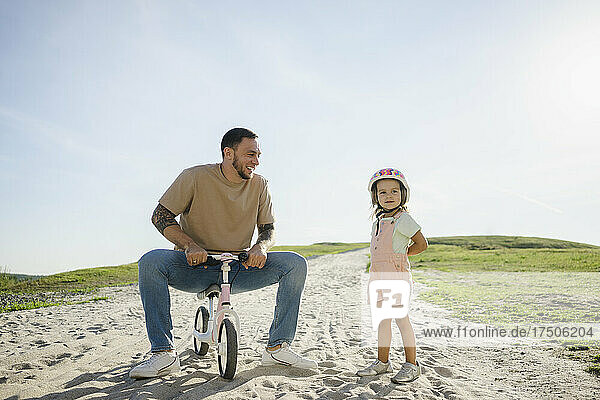 Playful father sitting on bicycle looking at daughter standing on sand