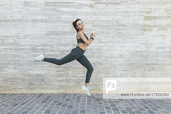 Smiling athlete jumping by wall on footpath