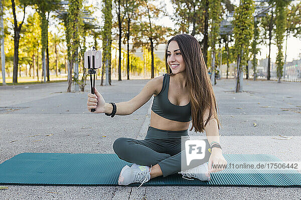 Smiling sportswoman with mobile phone vlogging on exercise mat in park