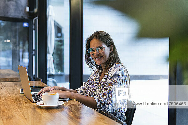 Smiling businesswoman with laptop sitting in coffee shop
