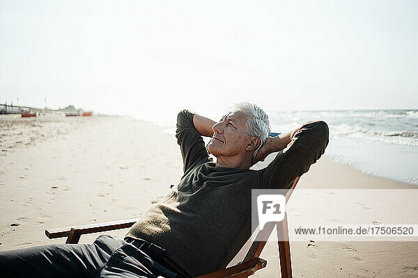 Älterer Mann mit den Händen hinter dem Kopf entspannt sich auf einem Stuhl am Strand