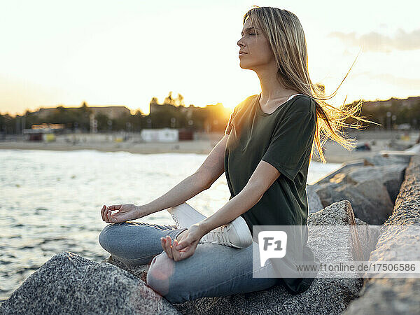 Blond woman meditating on rock at Bogatell beach  Barcelona  Catalonia  Spain