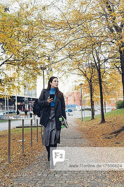 Businesswoman with smart phone walking on footpath in autumn