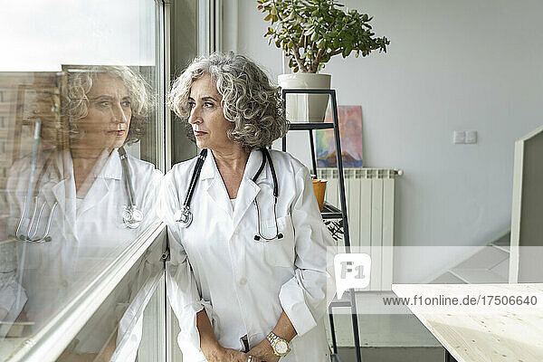 Thoughtful senior doctor looking out through window at home