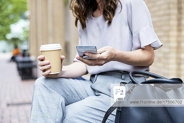 Geschäftsfrau sitzt mit Einwegbecher und Tragetasche auf einer Bank und nutzt ihr Smartphone