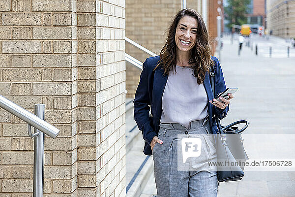 Smiling businesswoman with hand in pocket holding mobile phone walking on footpath