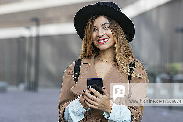 Woman wearing hat and trenchcoat holding smart phone in city