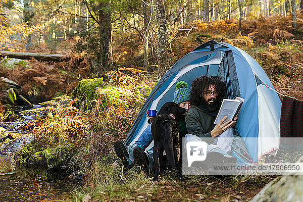 Hipster man reading book by girlfriend and dog in tent at forest