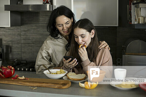 Mother and daughter looking at smart phone having breakfast in kitchen
