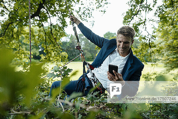 Lächelnder Geschäftsmann benutzt Smartphone  das im Park am Seil hängt