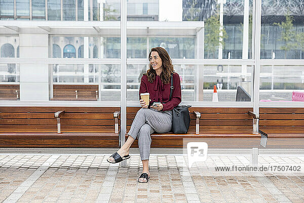 Businesswoman with disposable cup sitting on bench at railroad station