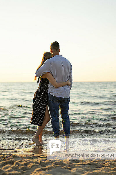 Romantic young couple with arms around looking at horizon from beach